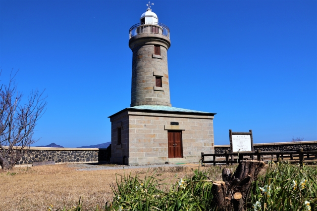青空の下、直島の草の生い茂った丘の上に、茶色の木製の扉がついた石造りの灯台が立っている。.