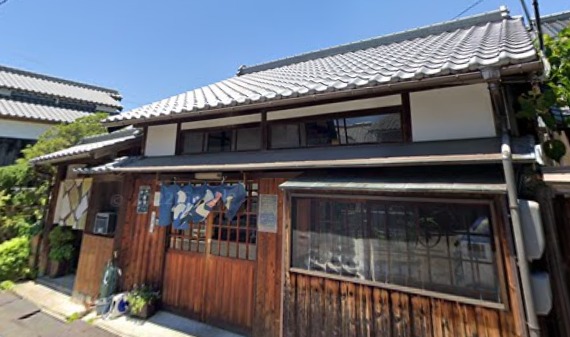 瓦屋根と木造のファサードを持ち、入り口には青いのれんが掛けられた伝統的な日本家屋。.